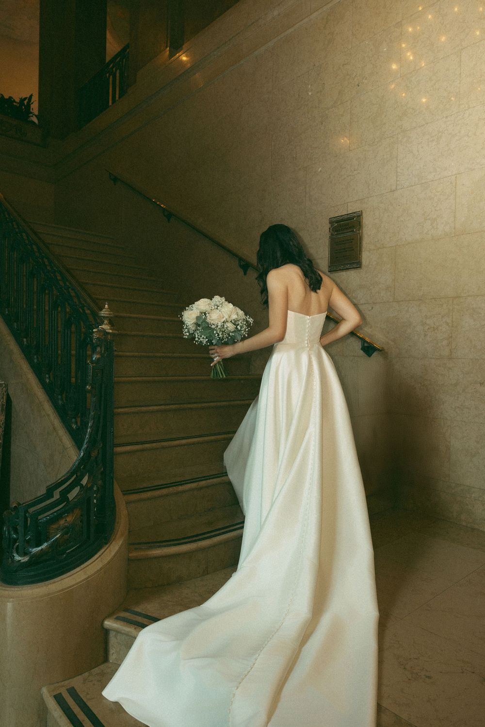 A bride in an elegant white strapless gown with long train ascends stone stairs while holding a bouquet.