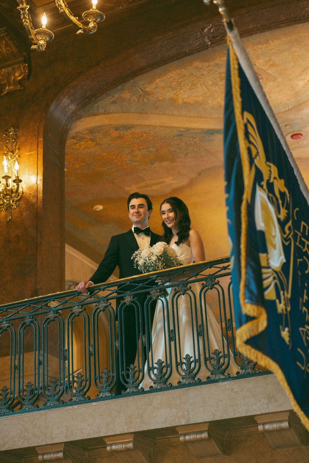Couple stands at an ornate balcony railing in classical interior with blue flags and golden wall sconces.