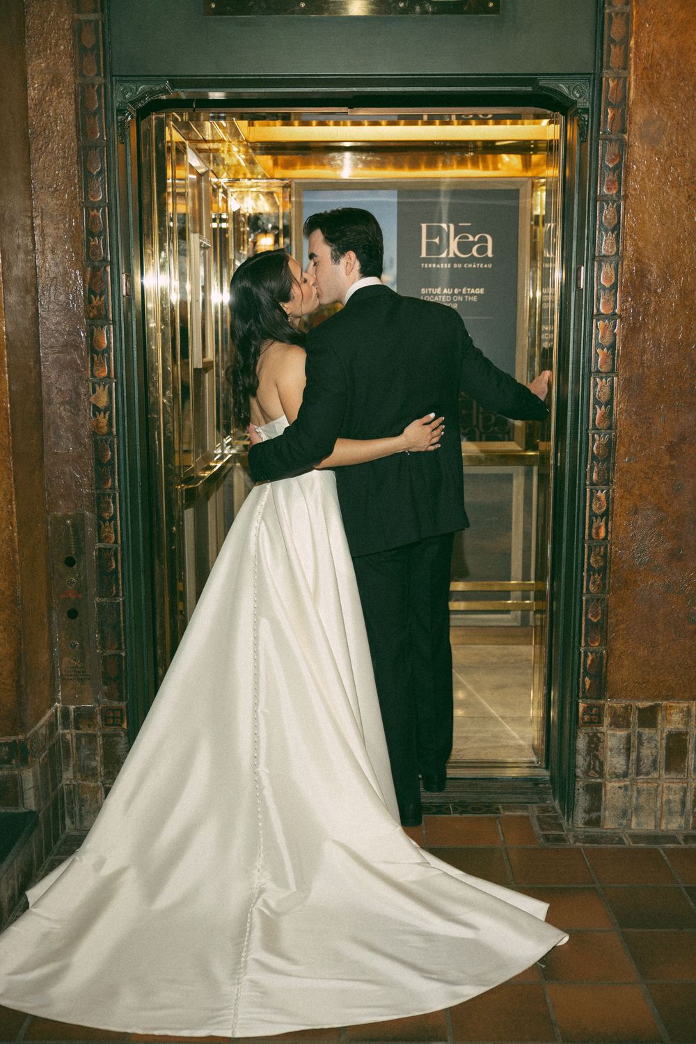 A newlywed couple shares a romantic kiss in an elegant elevator with ornate gold details and a flowing white wedding gown.
