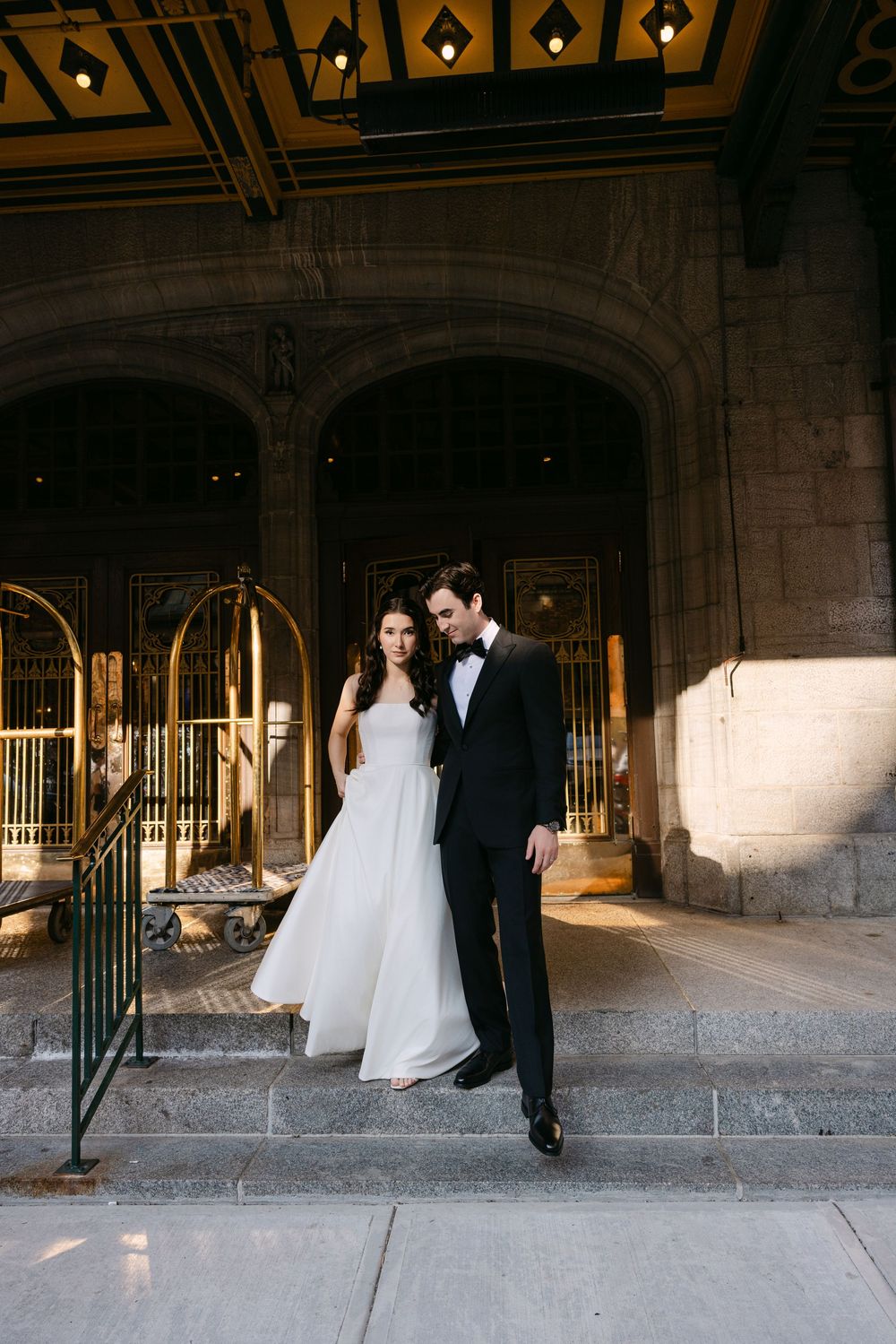 A wedding couple poses in formal attire beneath ornate gothic architecture with decorative arches.