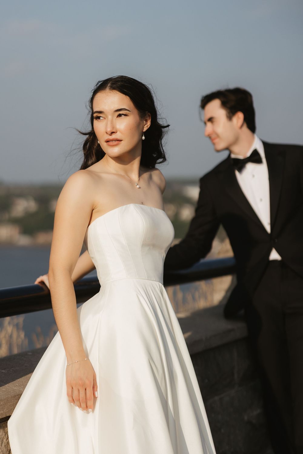 A couple in wedding attire poses against a waterfront backdrop, with the bride in a strapless white dress in the foreground.