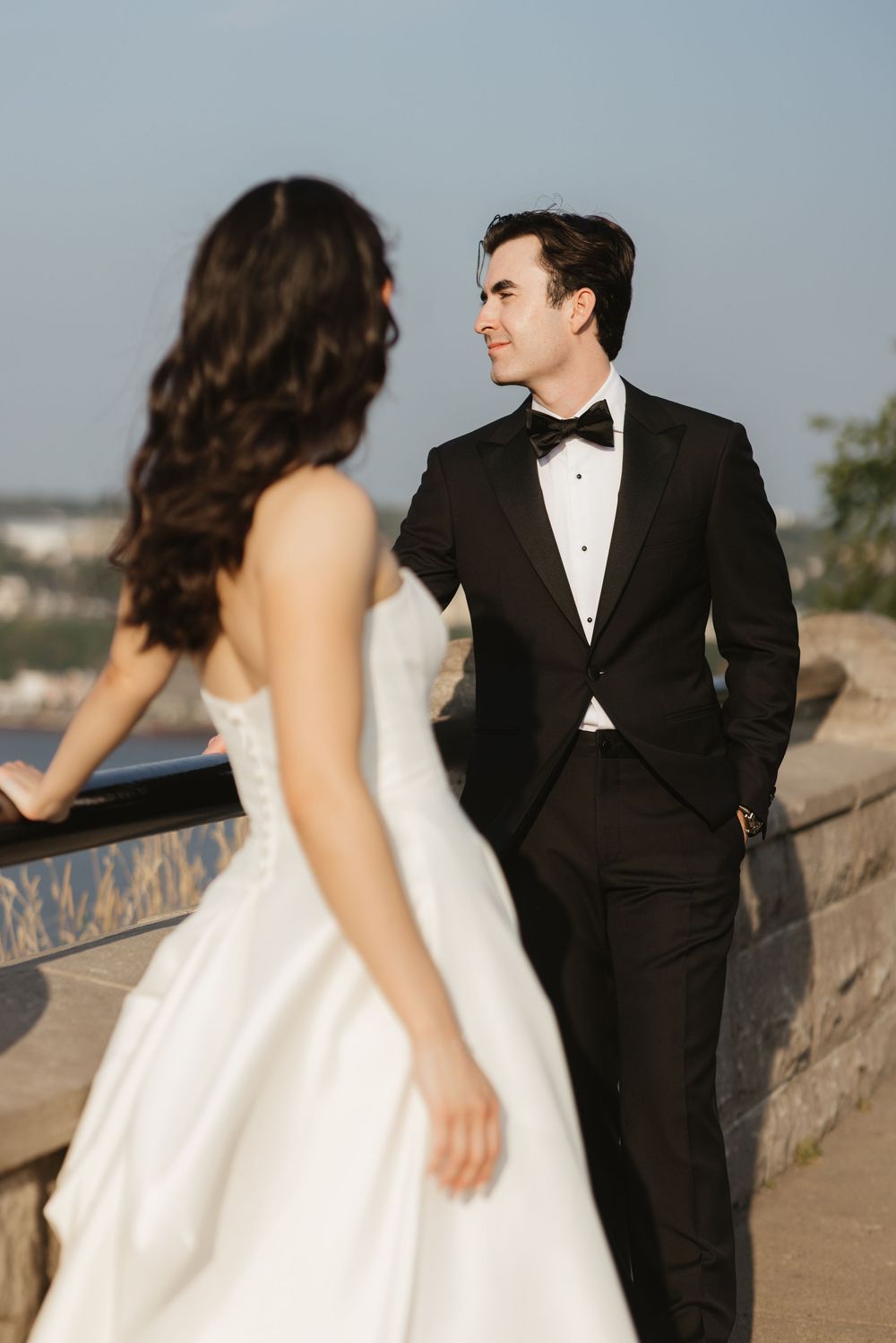 A couple in wedding attire share a moment on a stone wall overlook during their outdoor photoshoot.