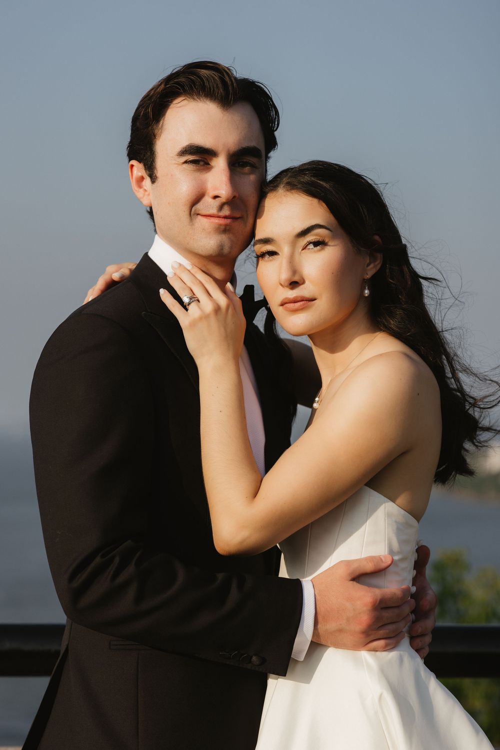 Bride and groom embrace in formal wedding attire against a sunset sky backdrop.