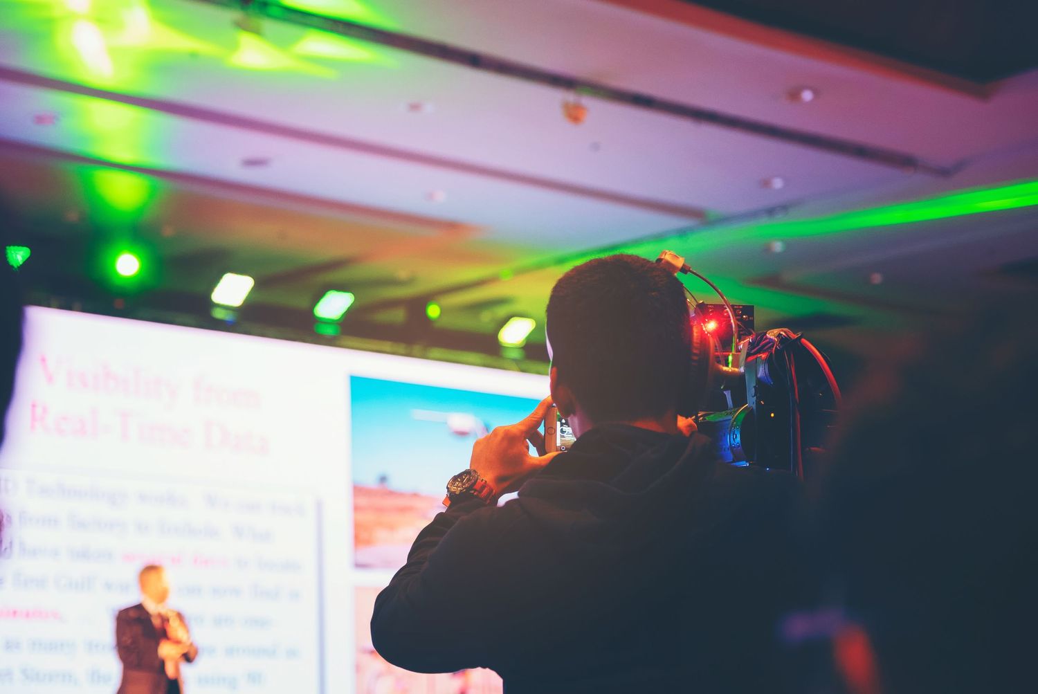 Concert photographer captures performance with camera while colorful stage lights illuminate the venue in green and pink hues.