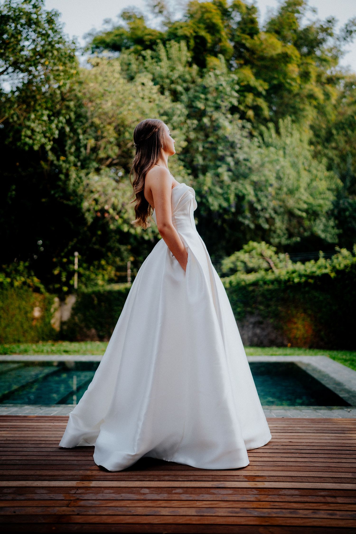 Flowing white wedding dress captured on wooden deck by pool with natural garden backdrop.