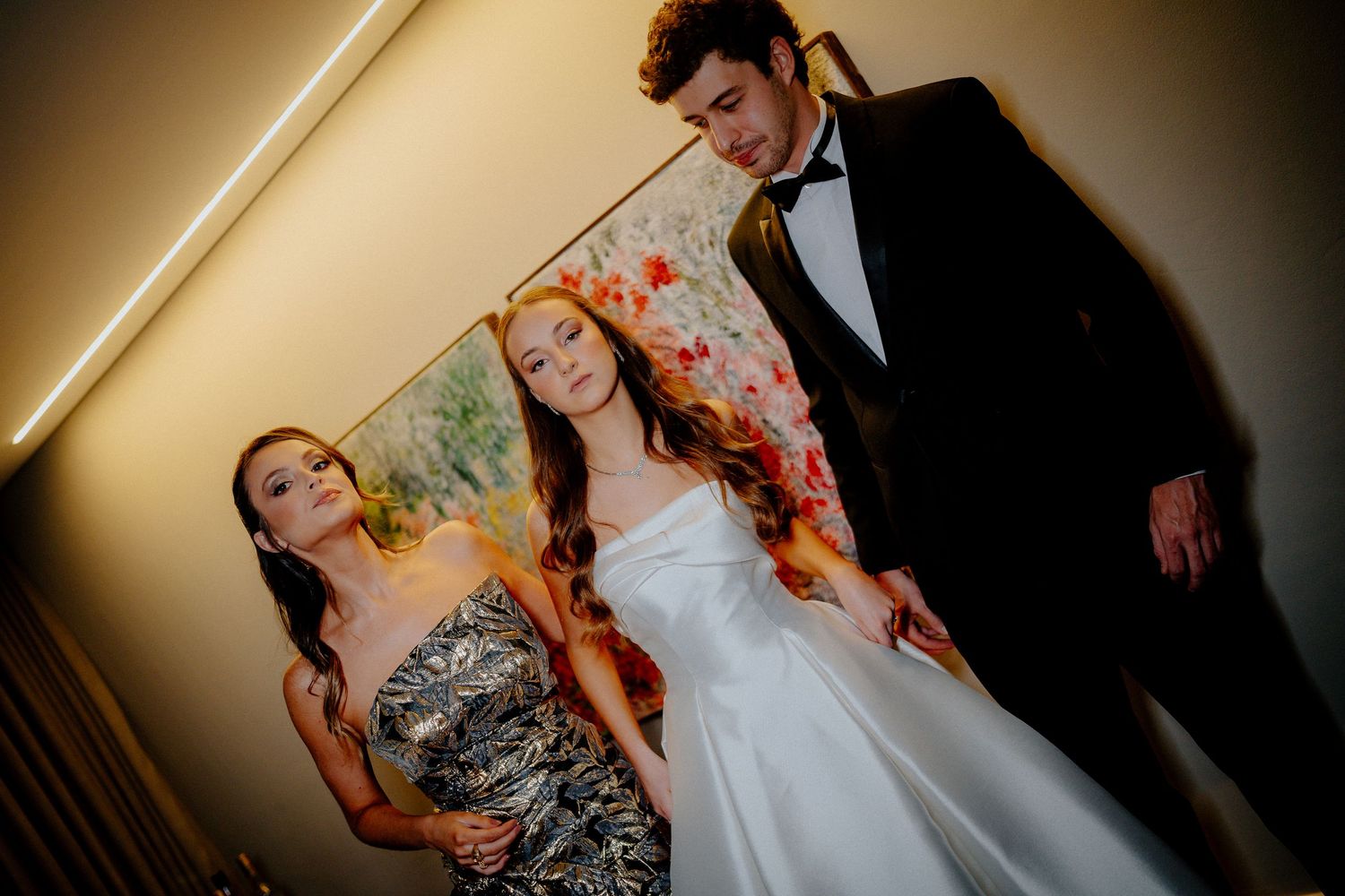 Three people in formal attire pose together against a wall with colorful floral artwork.