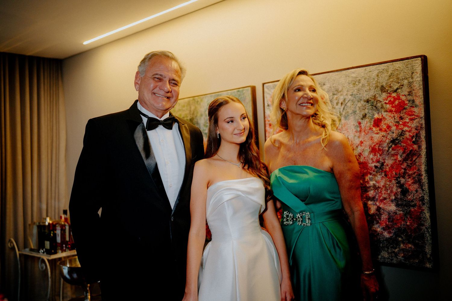 Three people in formal evening wear stand together against a wall with floral artwork.