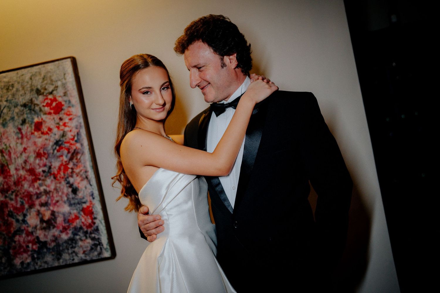 A couple embraces in formal attire against a wall with floral artwork.