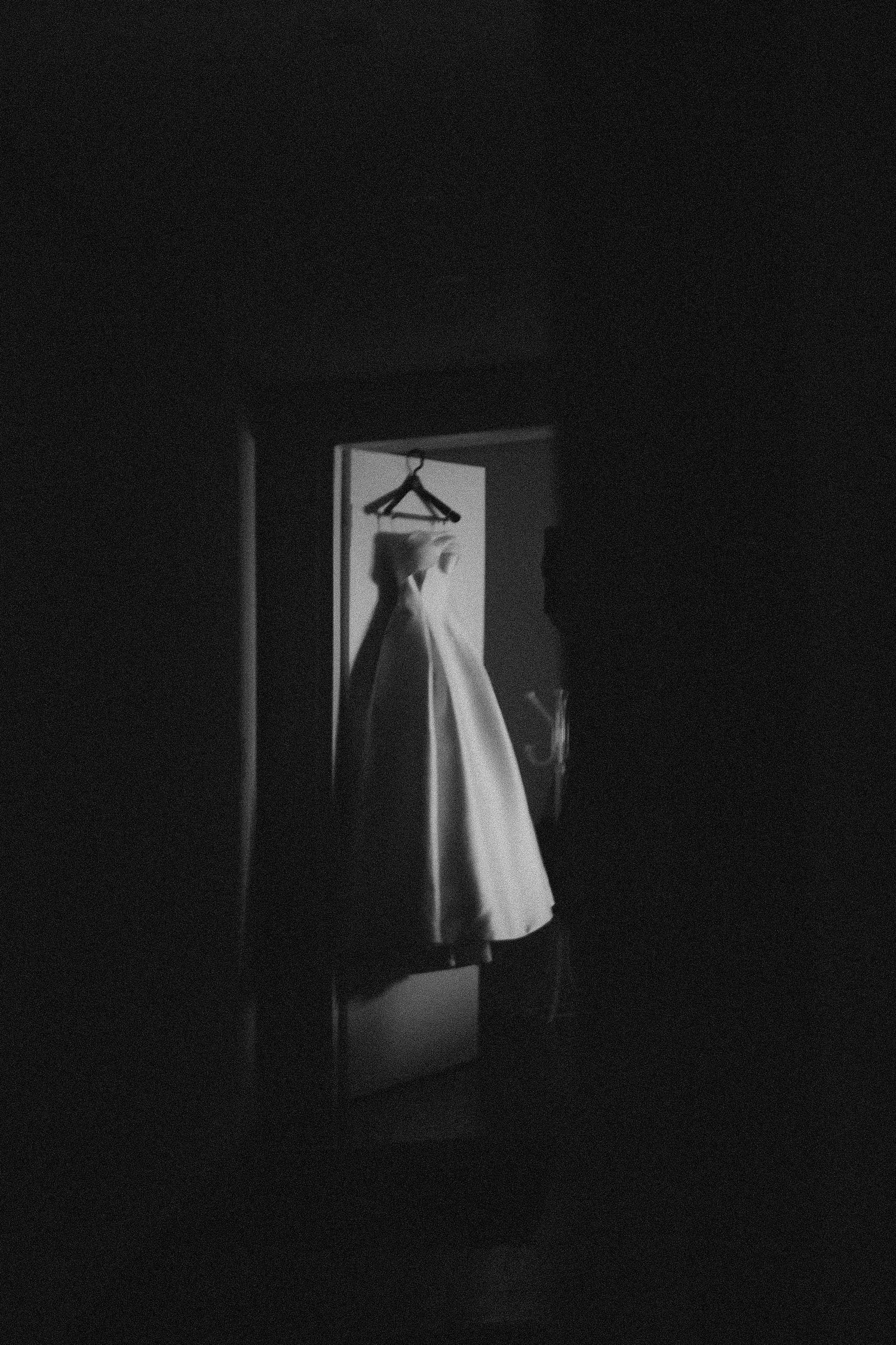 Classic white wedding dress hanging in doorway silhouetted against dark background.