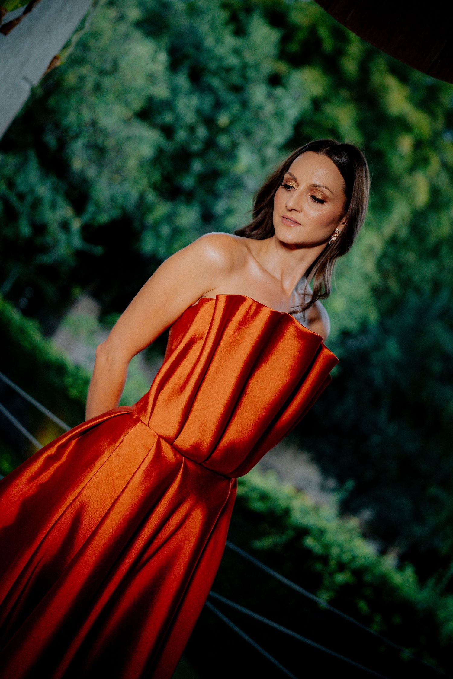 A woman in a stunning orange strapless gown poses outdoors against lush greenery.