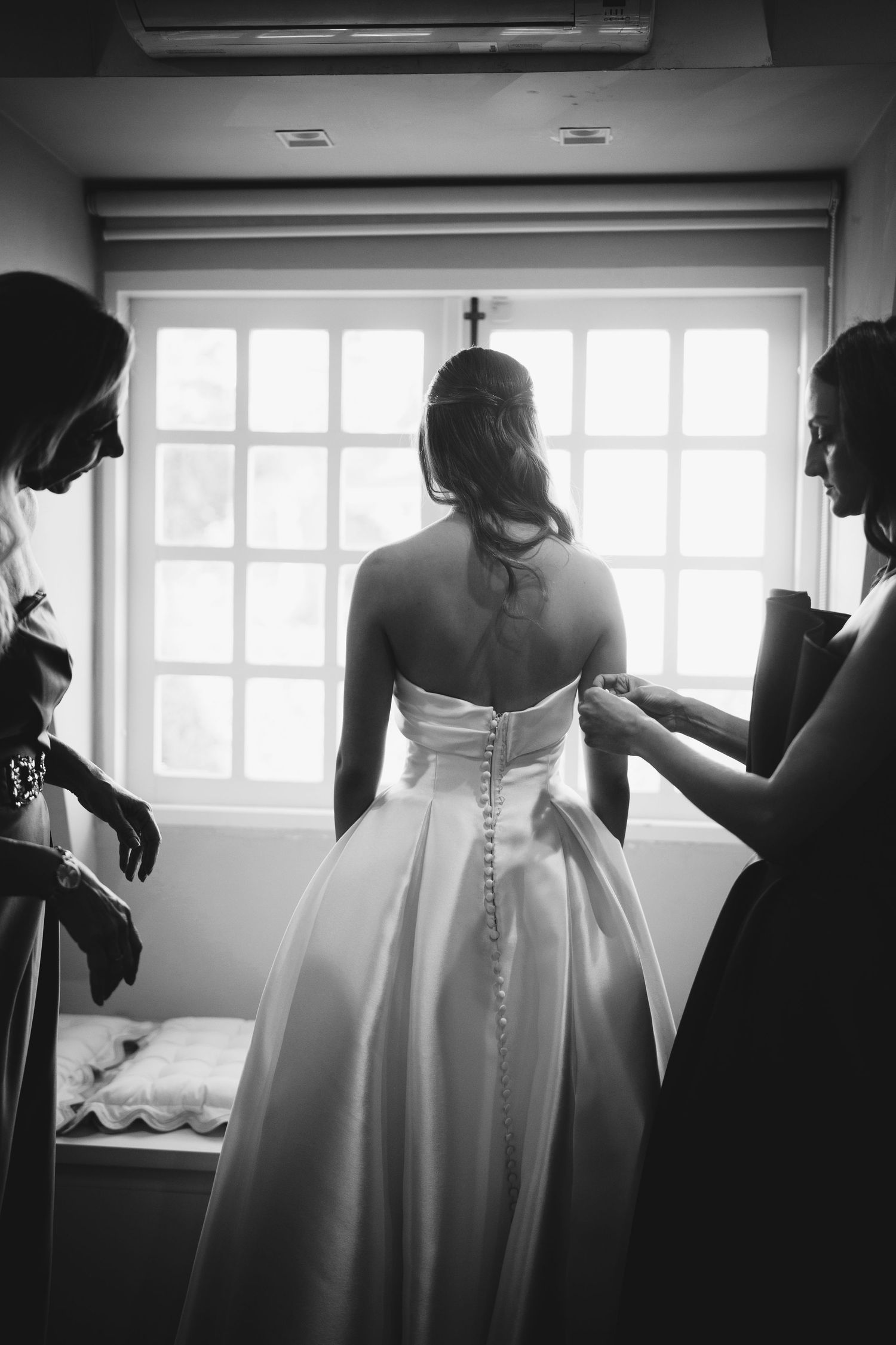 Classic wedding dress being buttoned up in front of bright window.