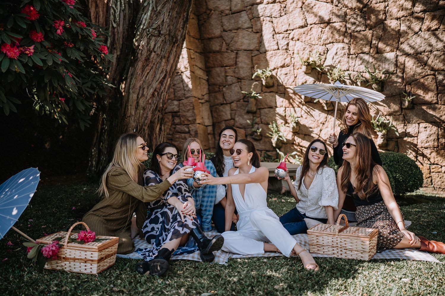 Group enjoying a sunny outdoor picnic with baskets and blankets on grass near stone wall.