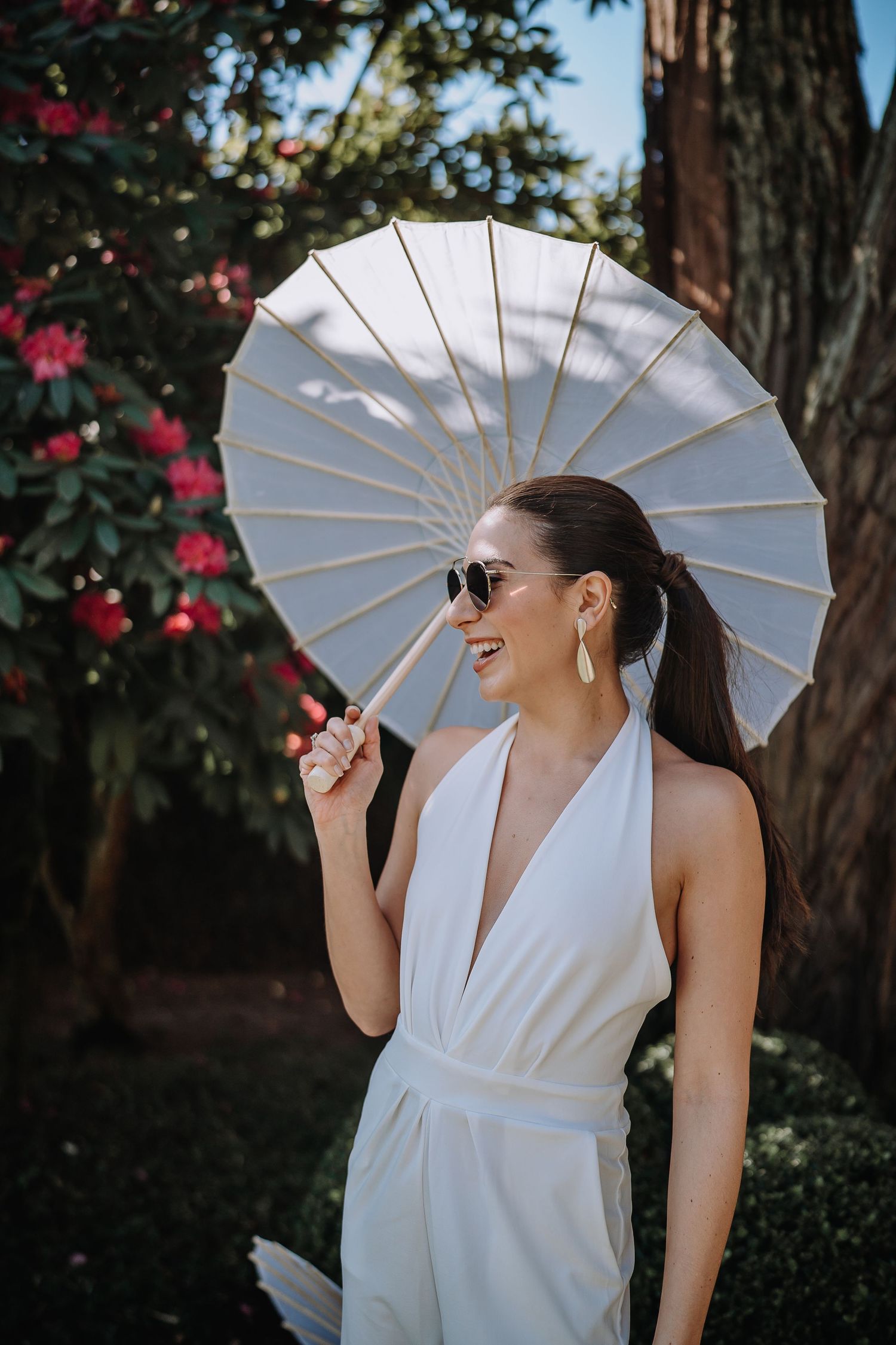 Woman in white jumpsuit holding parasol against flowering garden backdrop.