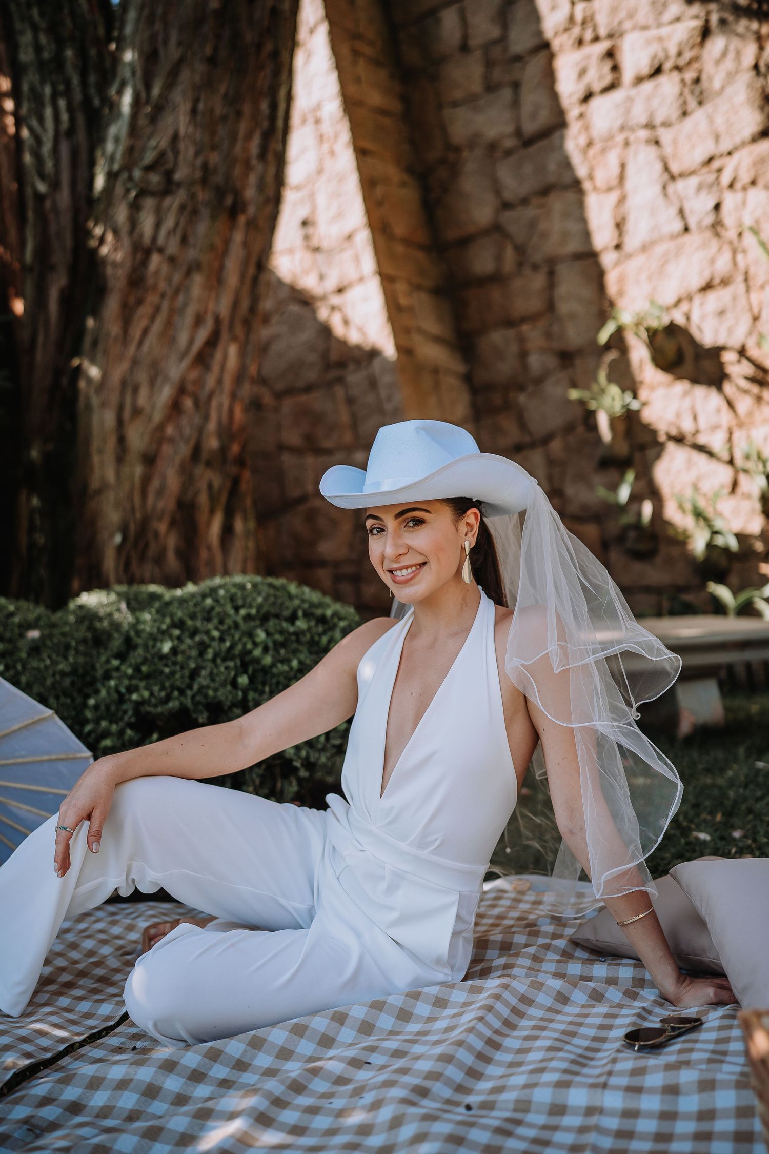 Woman in white jumpsuit and blue hat sitting on checkered blanket at outdoor celebration.