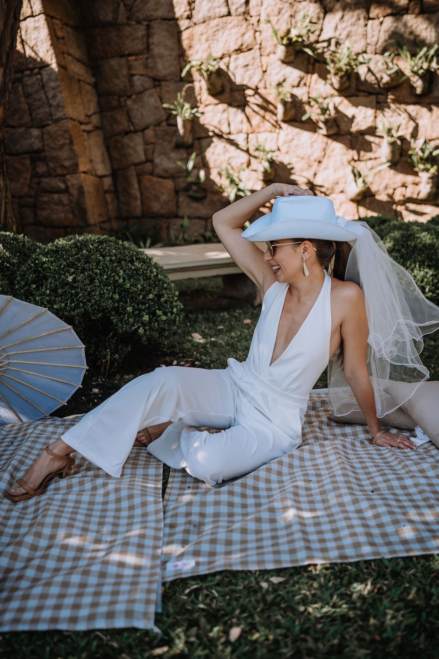 Person in white dress lounging on checkered picnic blanket in garden setting with stone wall backdrop.
