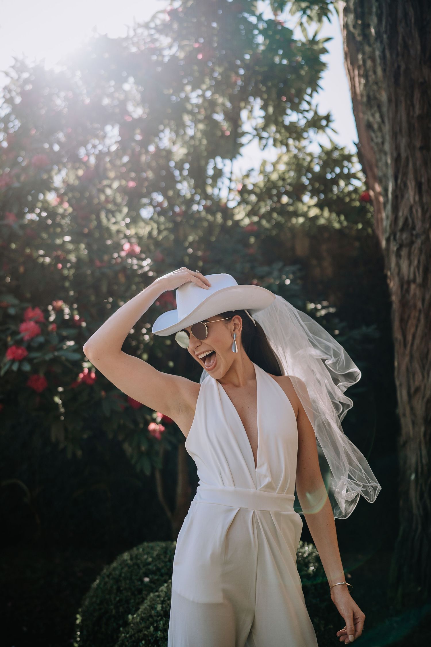 Figure in elegant white wedding dress poses against sunlit garden backdrop with trees and flowers.