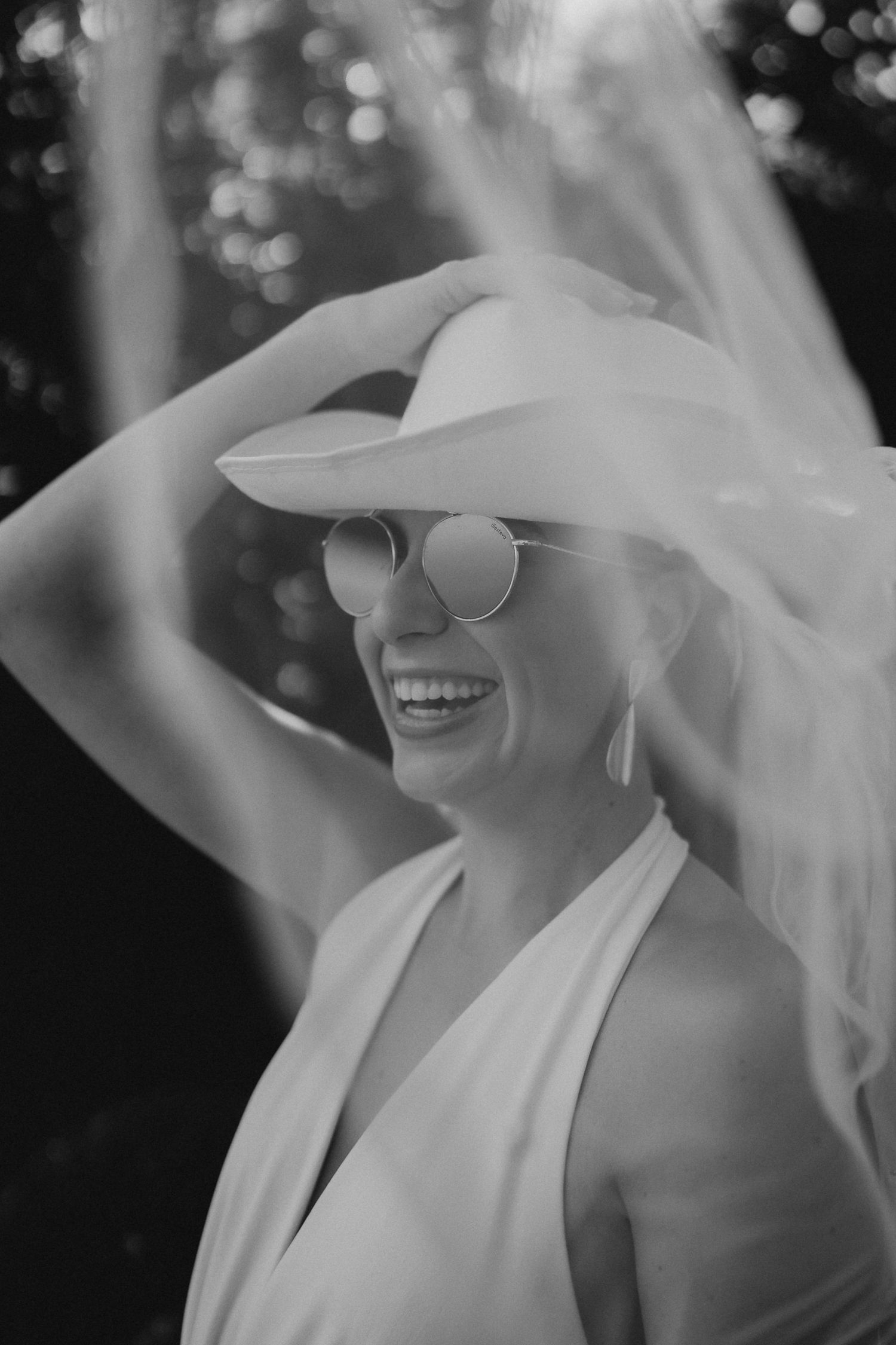 Black and white portrait of person in white dress and veil wearing sunglasses with joyful expression.