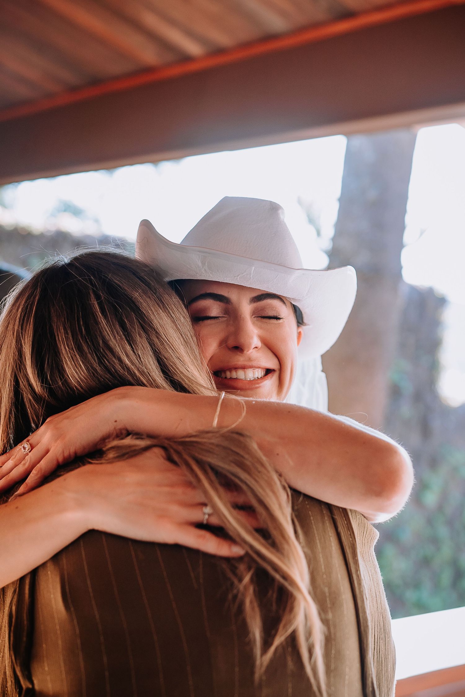 Warm embrace shared between people wearing white cowboy hats in sunlit setting.