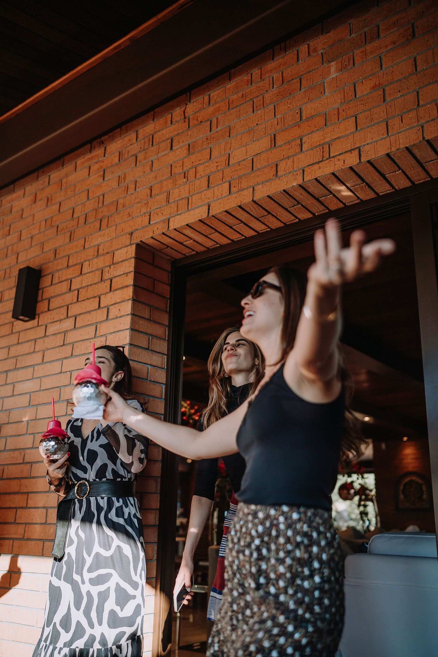 Friends dancing and celebrating with drinks at an outdoor party by a brick wall.