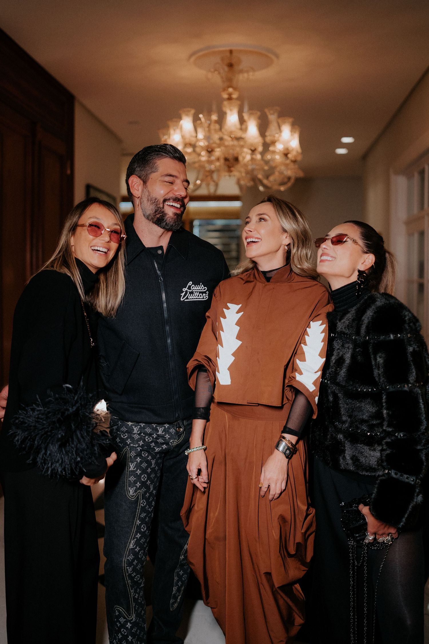 Friends in stylish winter outfits laughing together under chandelier at indoor gathering.