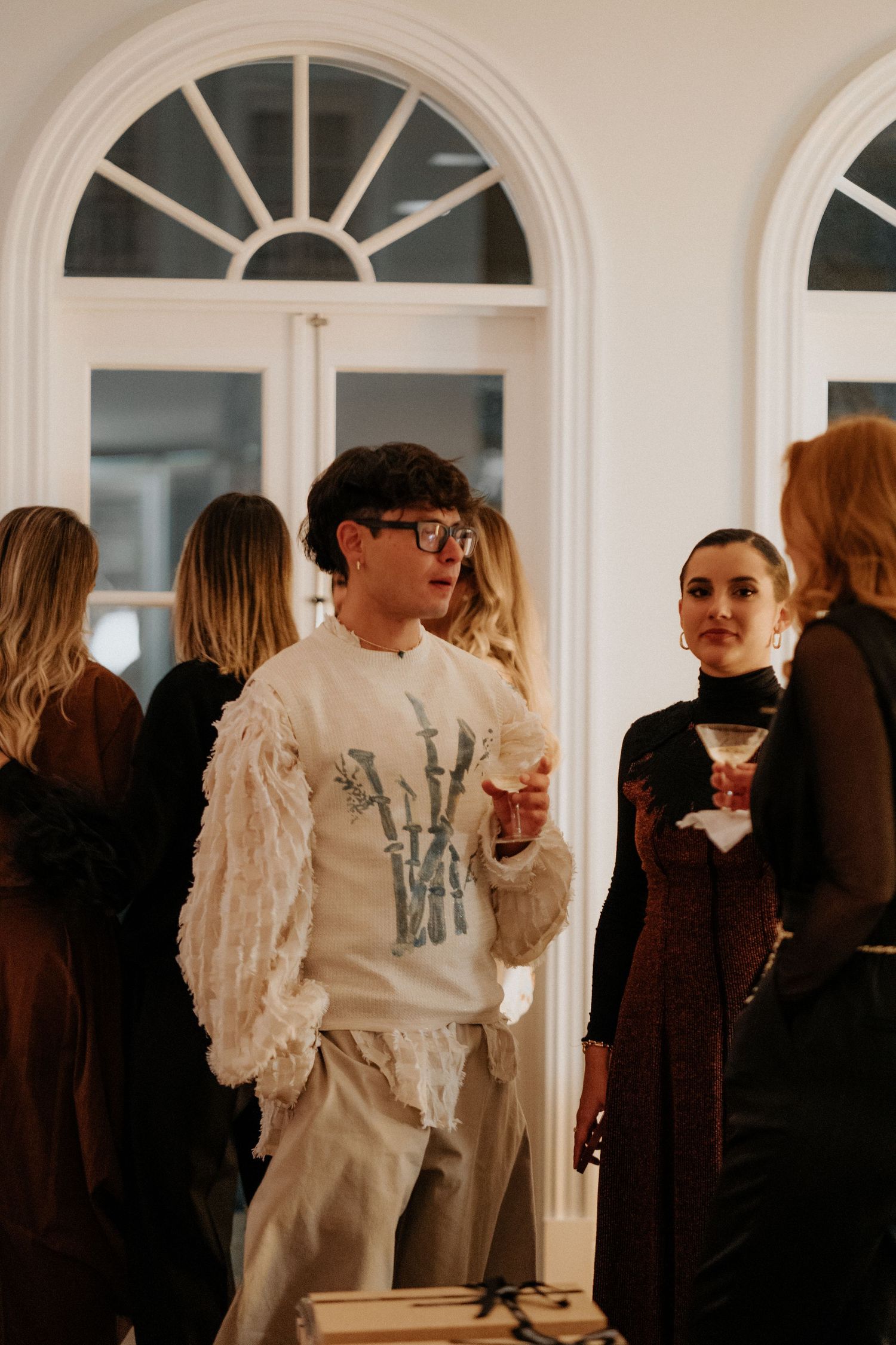 Elegant party scene with guests mingling near arched windows in formal setting.