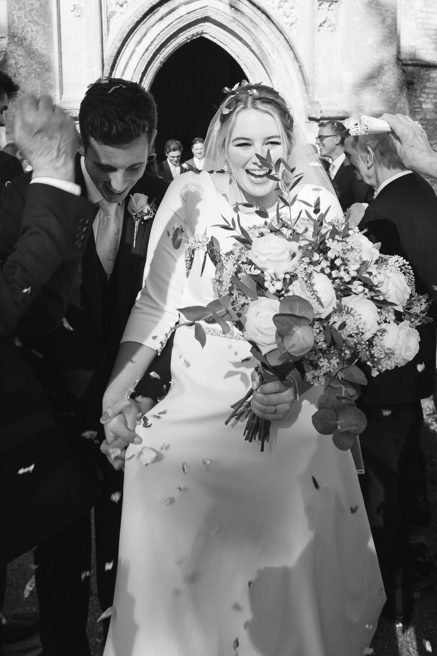Black and white wedding photo showing joyful moment outside church with bride holding beautiful floral bouquet.