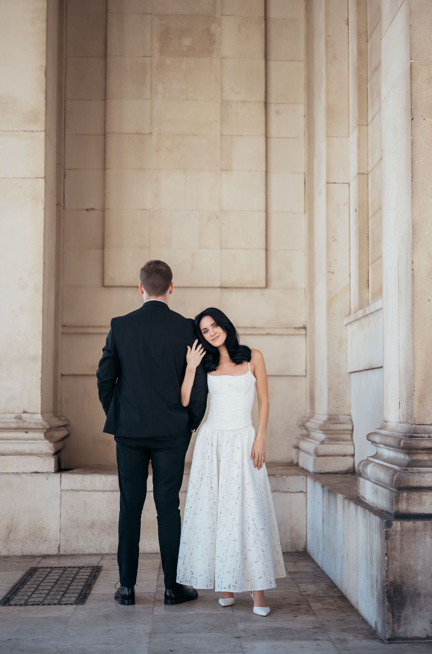 A couple in wedding attire stand between elegant stone columns in a classic architectural setting.