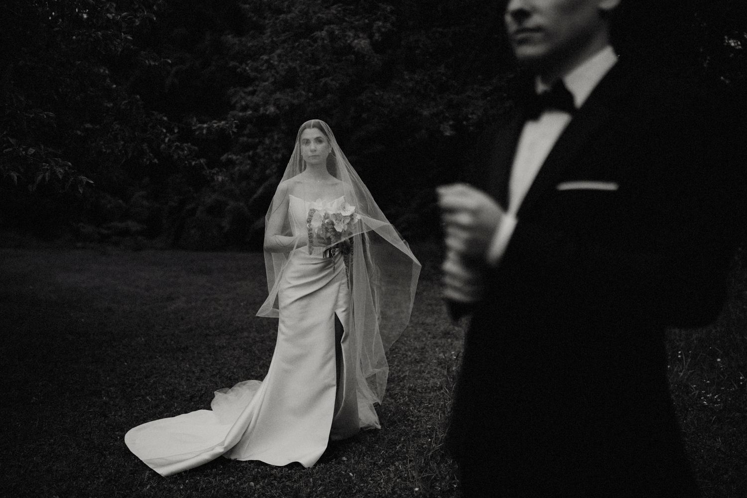 A black and white wedding photo showing a bride in a flowing dress and veil walking behind a suited figure in a garden setting.