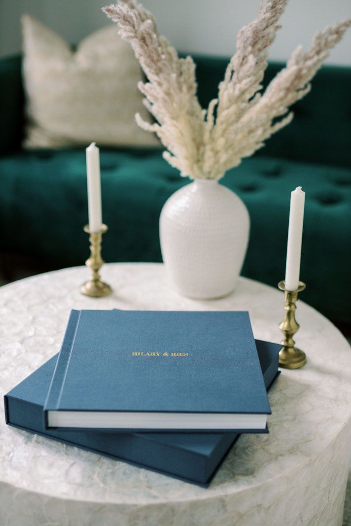Navy blue wedding photo albums with white pampas grass and candles on white textured surface.