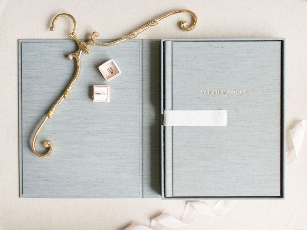 Elegant gray wedding album with decorative gold handle and two delicate diamond rings on light background.