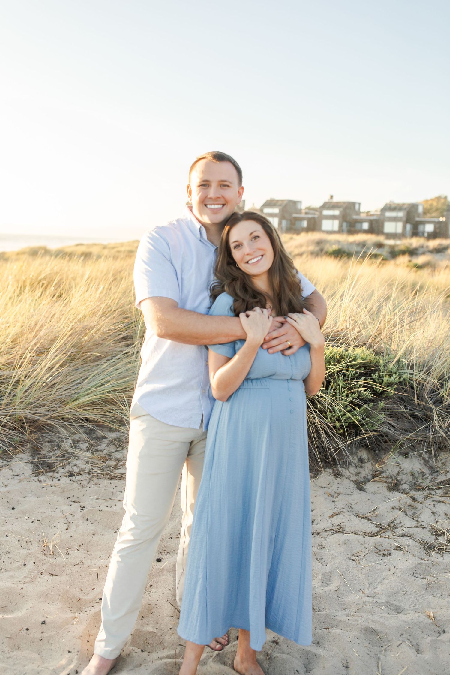 A couple embraces on a sandy beach near coastal homes at sunset with tall grass in the background.