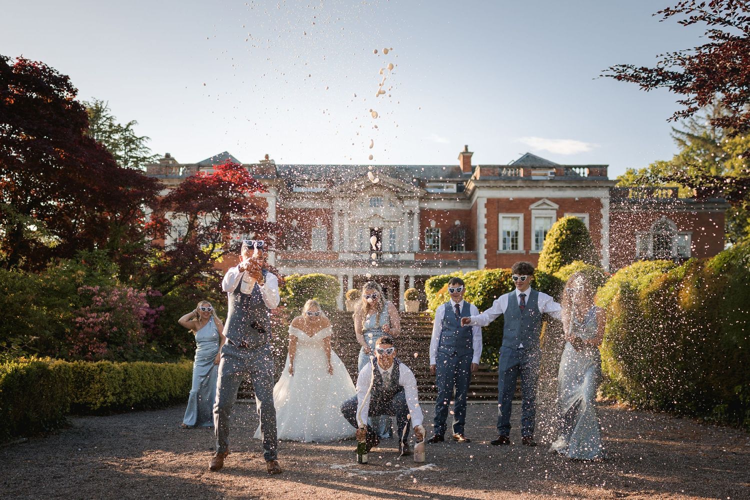 Wedding party celebrates with sparklers at sunset in front of historic brick mansion during outdoor reception gathering.