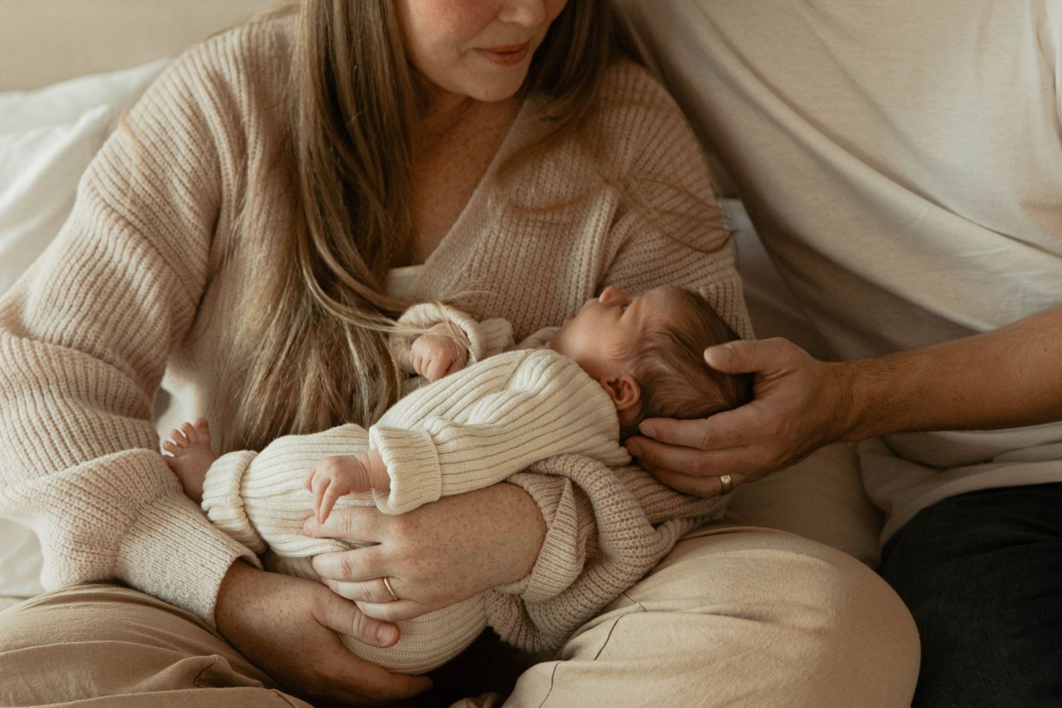 Parent in beige sweater cradling newborn baby wearing white knit outfit.