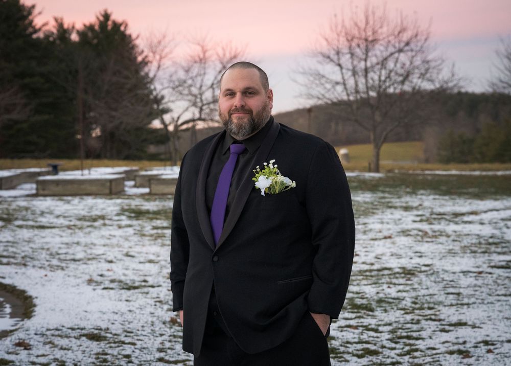 Someone in a black suit with purple tie and boutonniere stands in a snowy field at dusk.