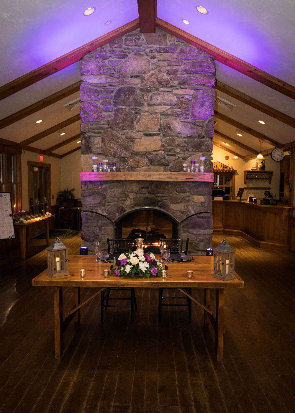 Rustic reception venue with stone fireplace, wooden floors, and purple uplighting creating a warm ambiance.
