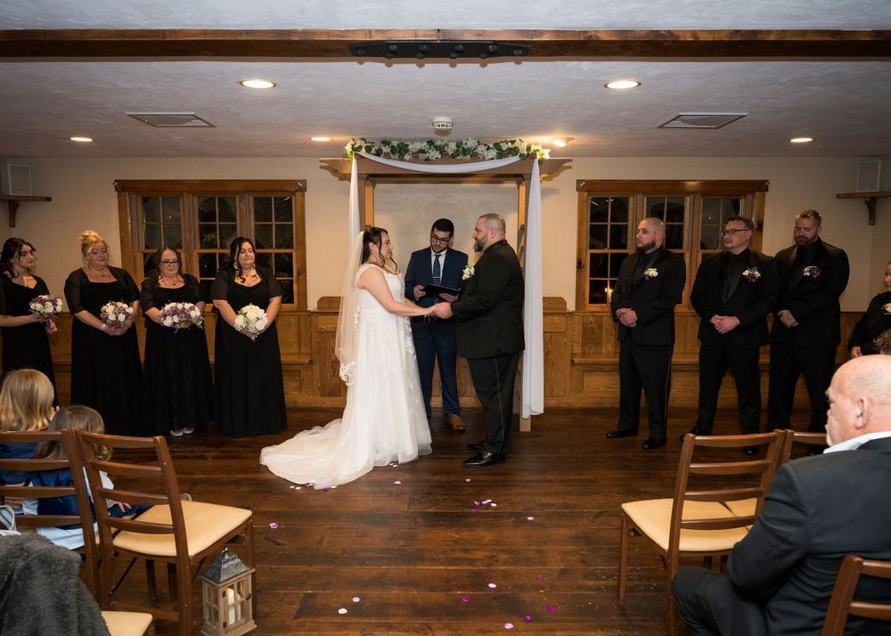 Indoor wedding ceremony with wedding party in black and white attire standing in front of a decorated arch.