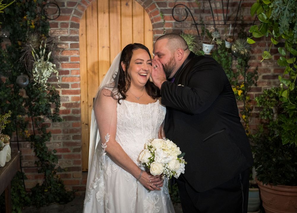 A couple in wedding attire share intimate moments against a rustic brick wall backdrop with greenery.