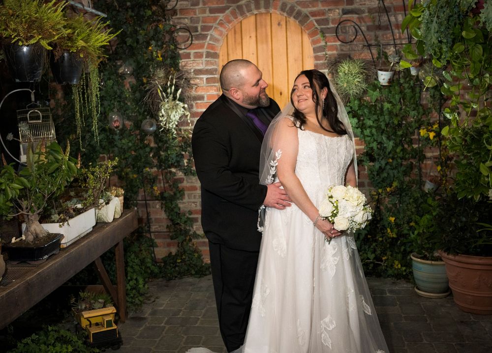 A couple shares an intimate moment in a romantic garden setting with brick walls and lush greenery surrounding them.