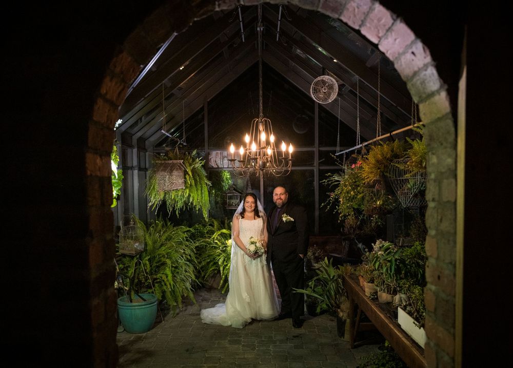 Newlyweds pose under a rustic wooden archway decorated with greenery and chandeliers at night.