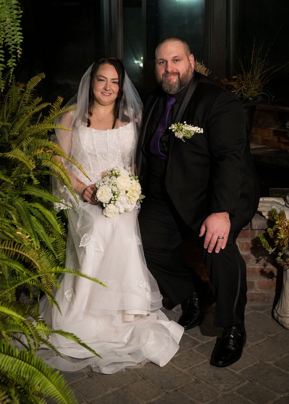 A wedding couple sits together on stone steps at night surrounded by lush greenery and soft lighting.
