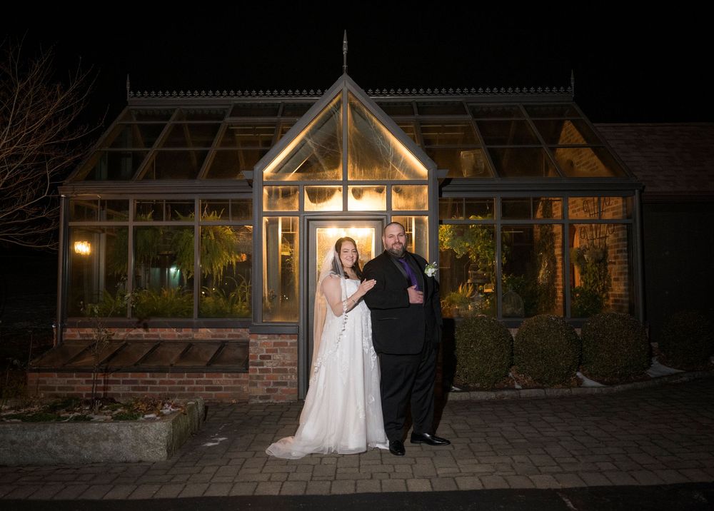 A couple in formal wedding attire stands in front of a rustic wooden building illuminated by warm evening lights.