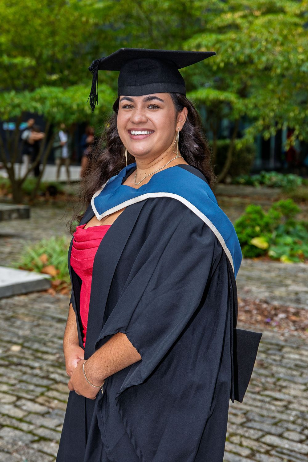 Graduate in black cap and gown poses outdoors on graduation day with bright greenery in background.