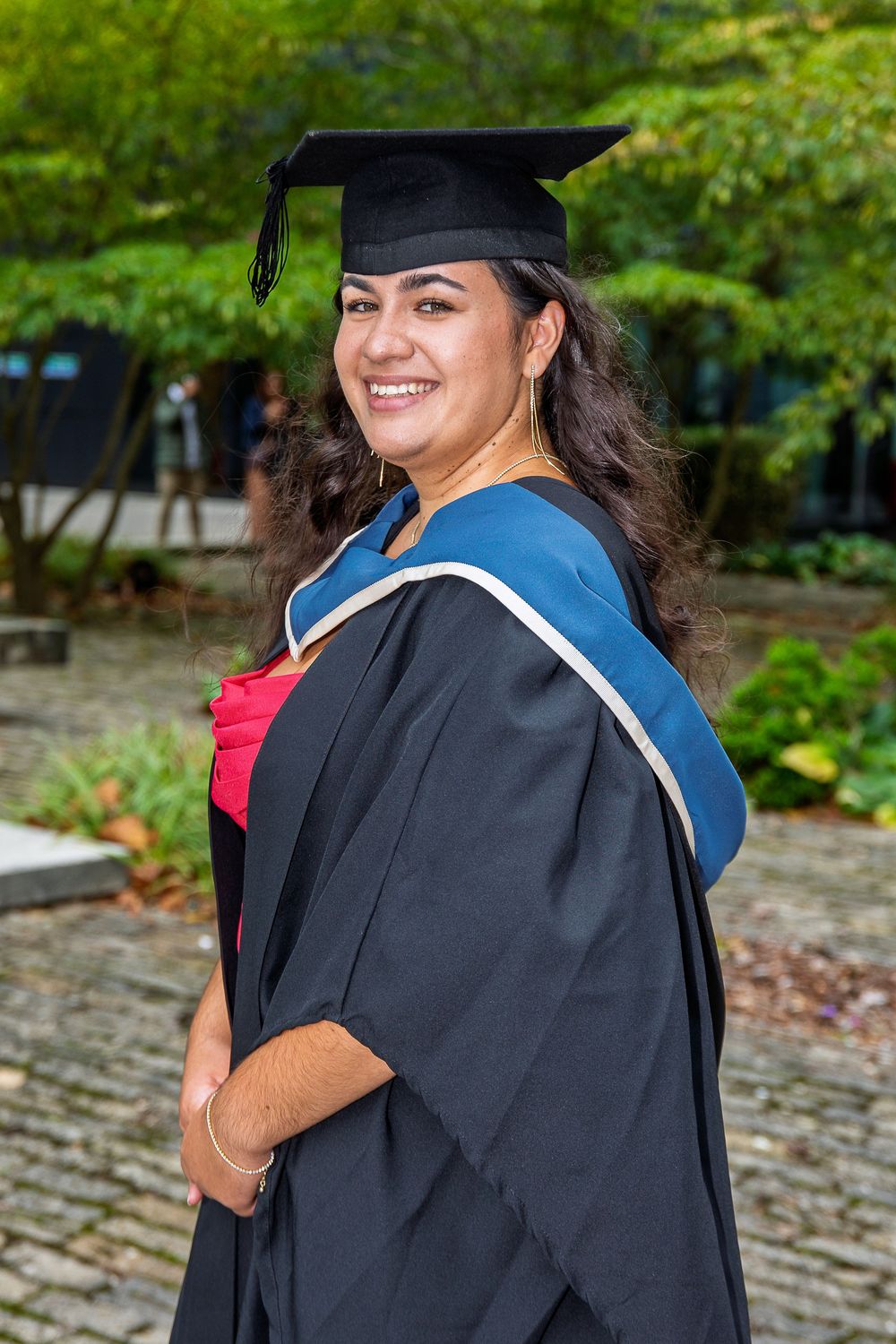 Graduate in traditional academic regalia smiles while posing against green outdoor backdrop.