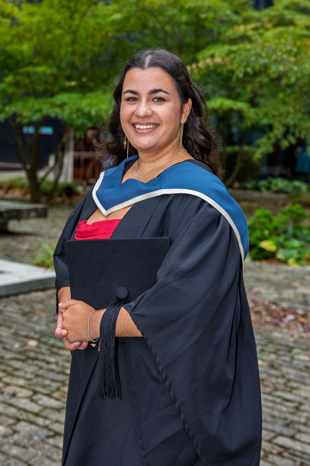 Graduate wearing academic regalia smiles while posing in front of lush garden setting.