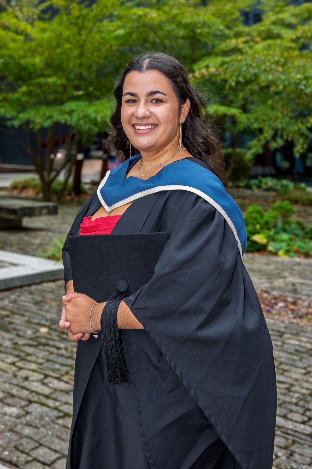Graduate wearing black academic regalia smiles during outdoor commencement ceremony on a sunny day.
