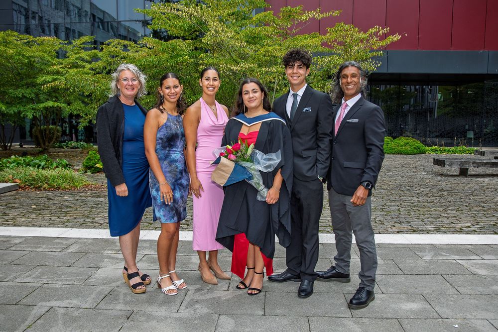 Extended family gathering for graduation photos outdoors with greenery and buildings in background.
