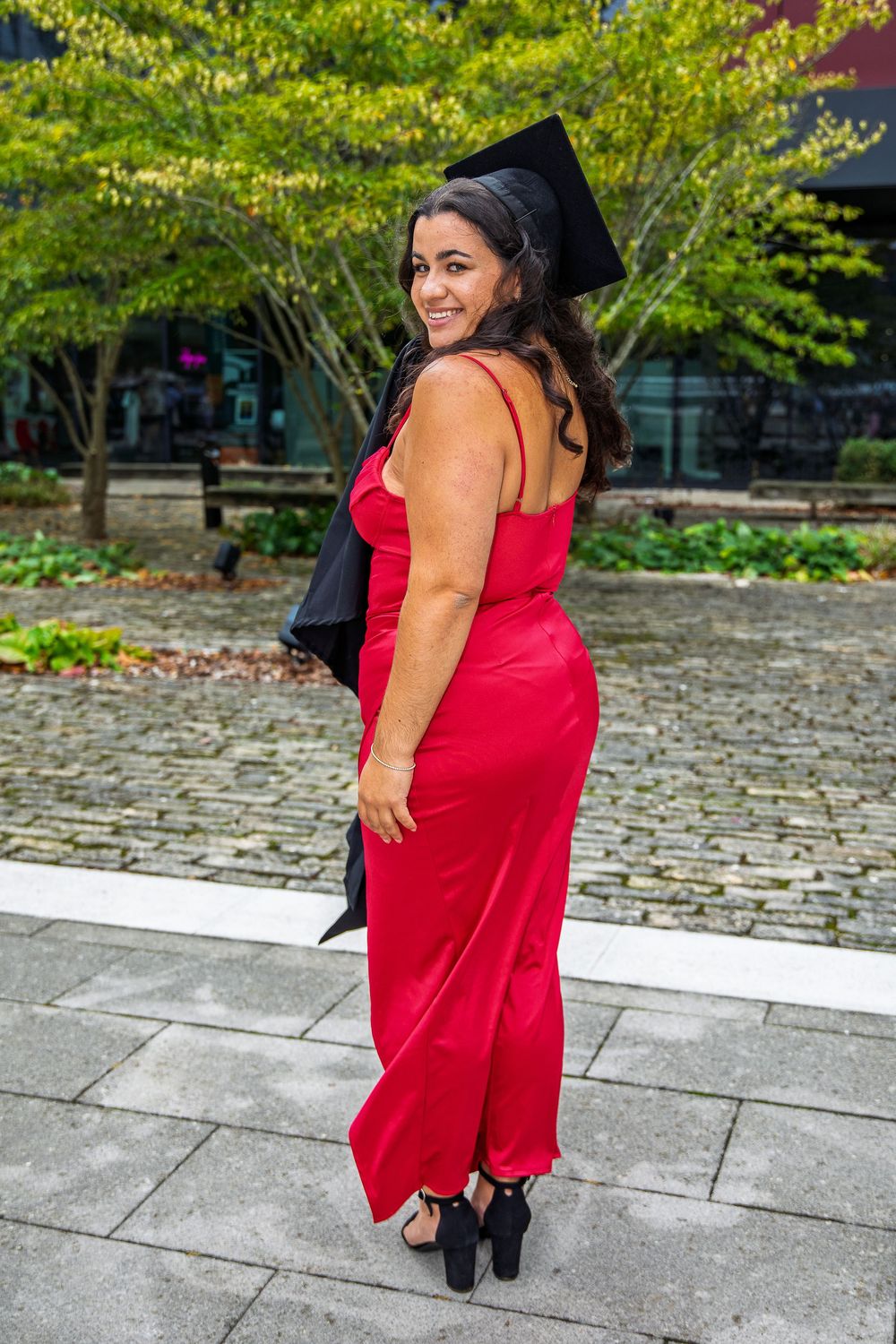 Graduate in red dress and black cap and gown poses outdoors during commencement.