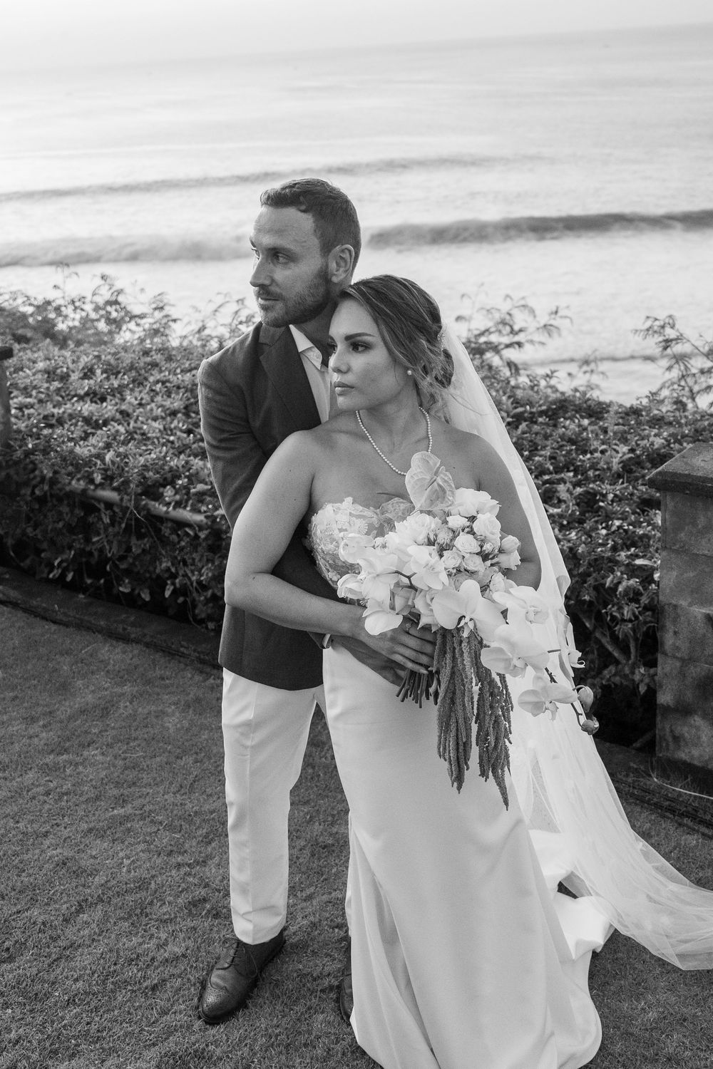 A tender black and white beachside wedding moment with a couple embracing while holding an elegant white bouquet.