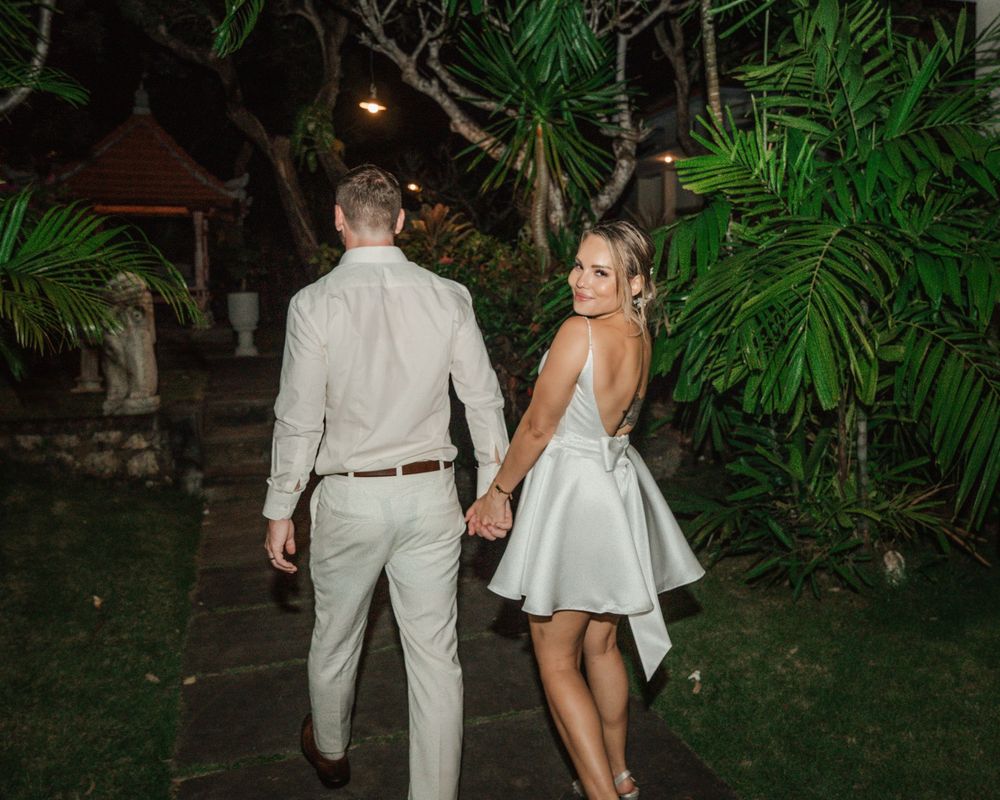 A couple holding hands walk through a tropical garden at night wearing formal light-colored attire.