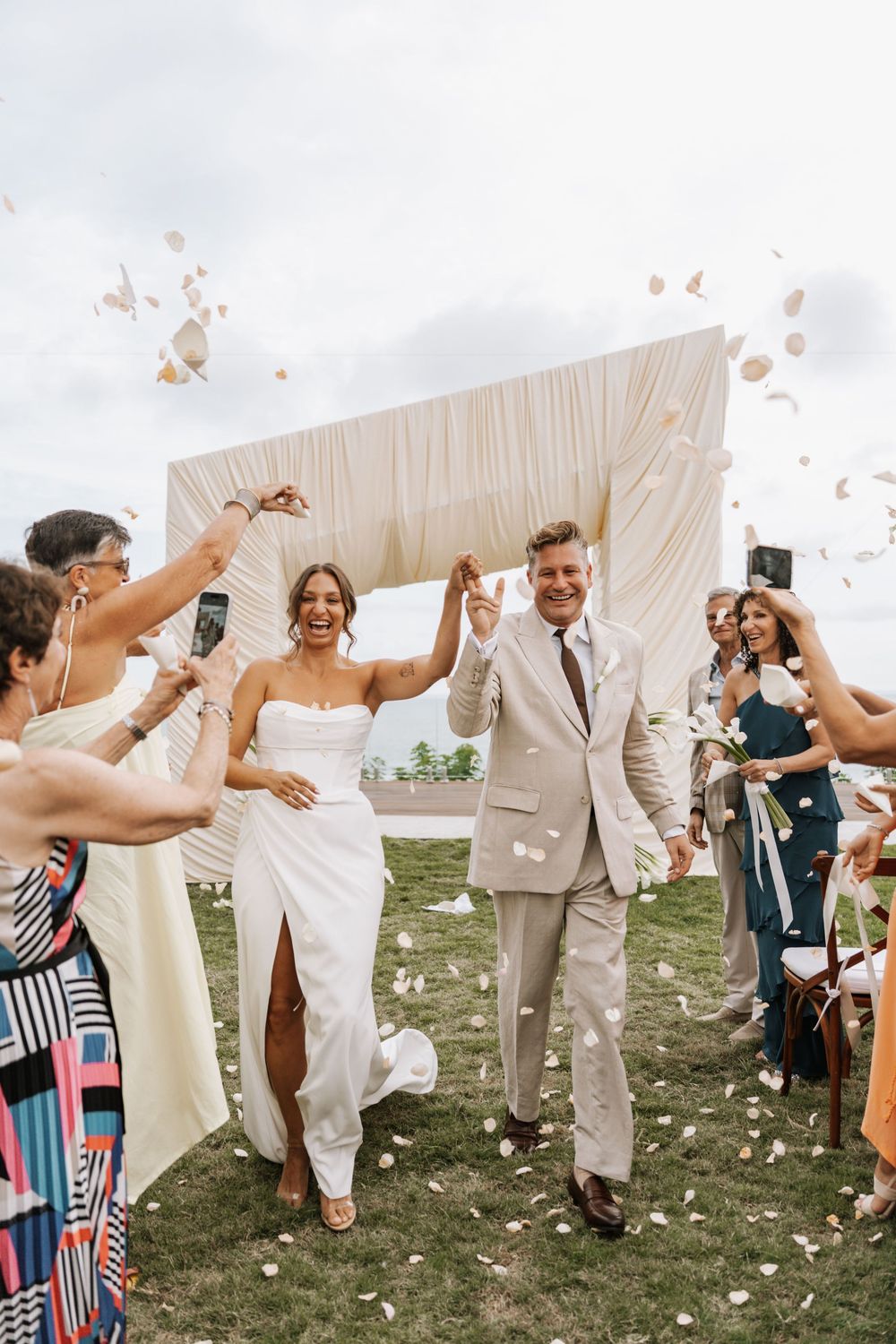 Wedding guests celebrate with flower petals and confetti outside under a white tent on a sunny day.